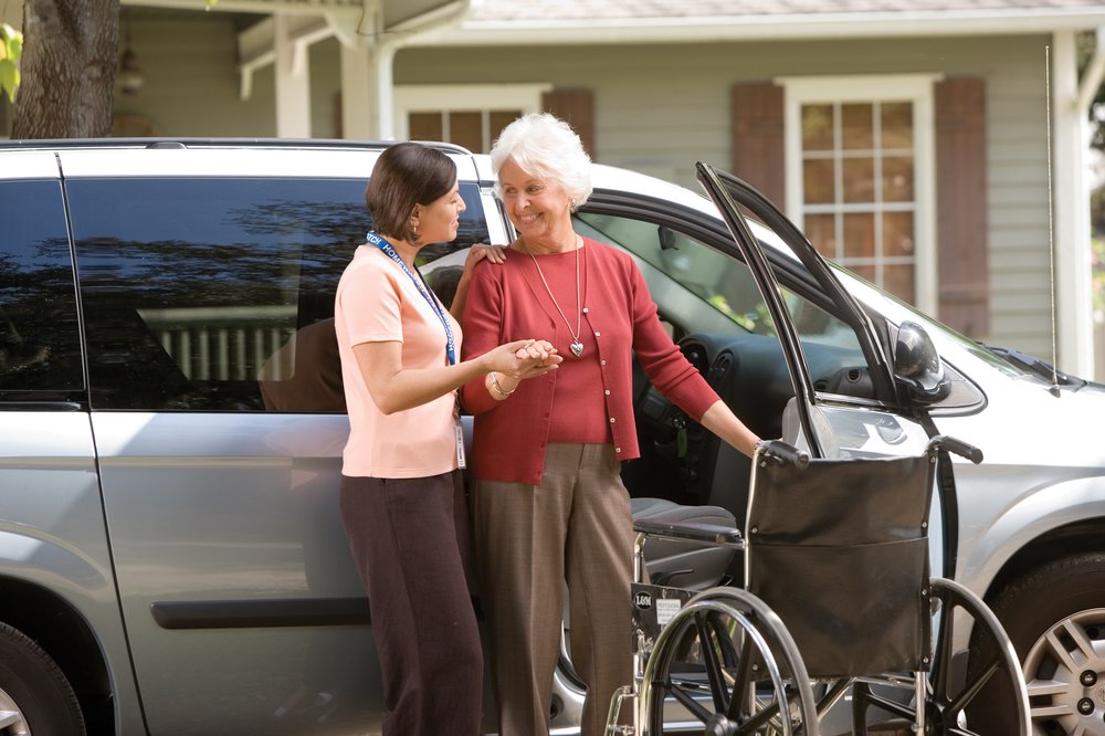 A female caregiver helping a senior woman from her wheelchair to her car.